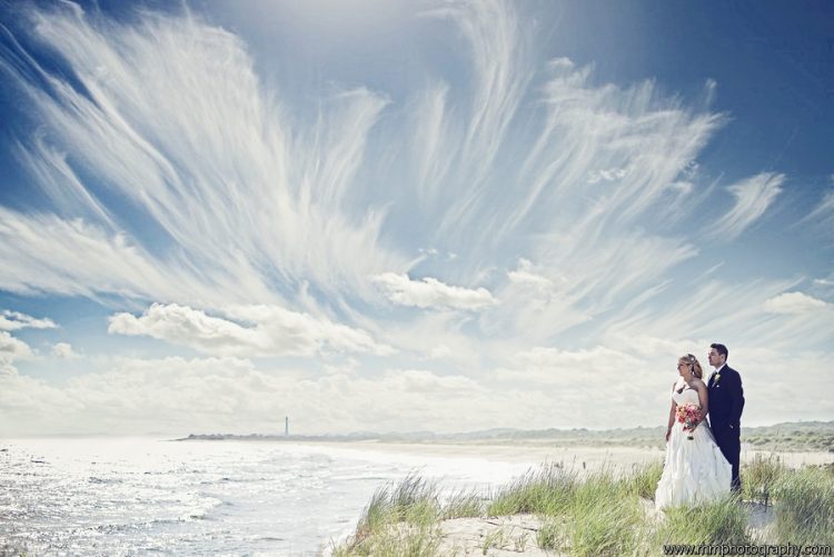 Bride and groom standing together on beach beneath dramatic cloud-filled sky during wedding portrait session