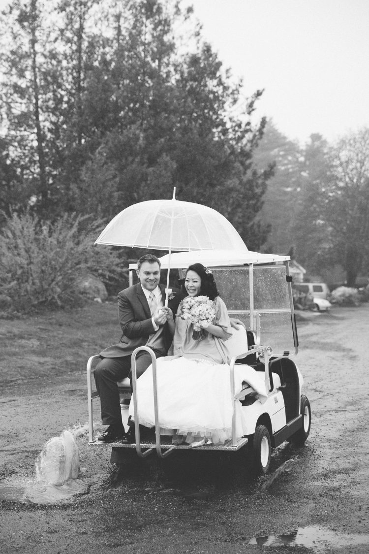 Bride and groom riding in golf cart under umbrella in rainy outdoor setting