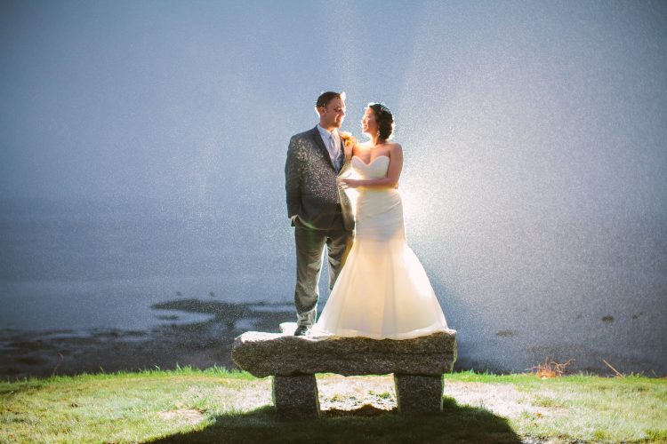 Bride and groom standing on stone bench by waterfront with sunlight behind