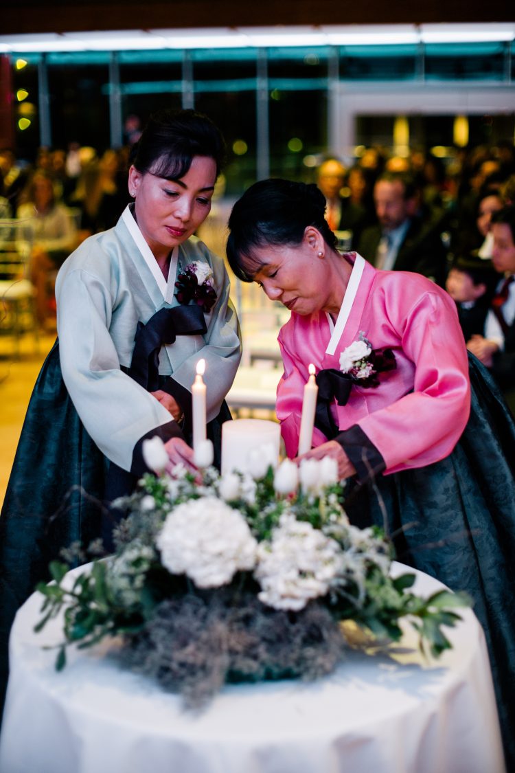 Two women in traditional Korean hanbok attire arranging candles and floral centerpiece during wedding ceremony reception