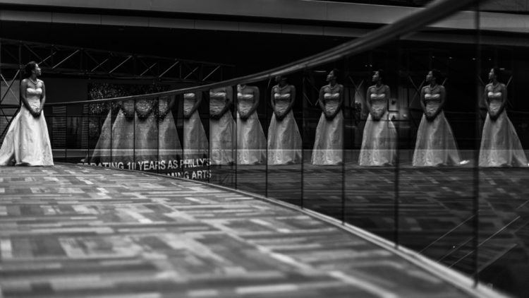 Black-and-white artistic reflection portrait of bride standing alone in elegant wedding gown beside mirrored wall