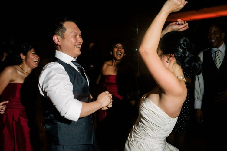 Bride and groom dancing joyfully together during lively wedding reception celebration with cheering guests nearby