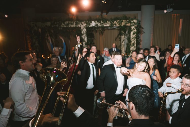 Packed wedding dance floor with brass musicians and guests dancing around bride and groom.