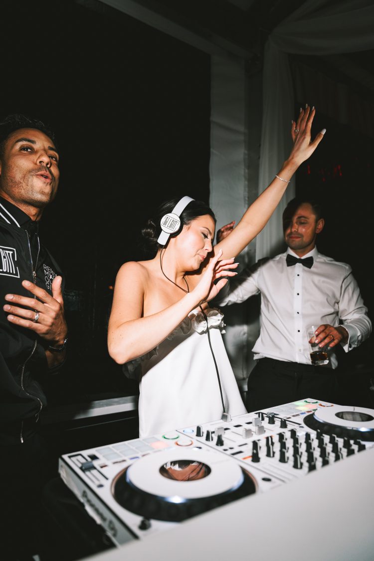 Bride DJing during wedding reception while Groom dances beside the DJ booth.