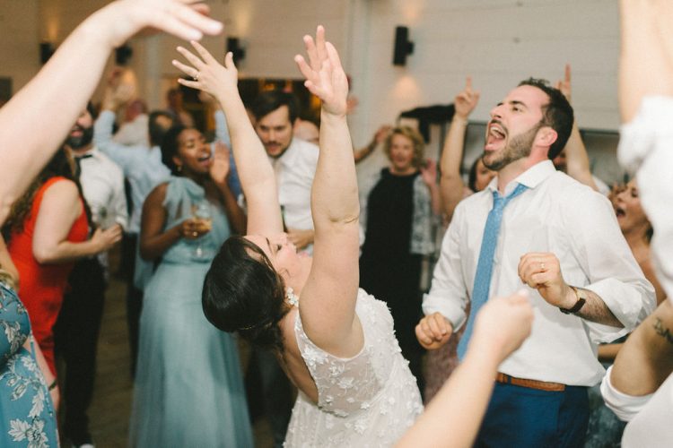 Bride dancing with raised arms while guests sing along on dance floor.