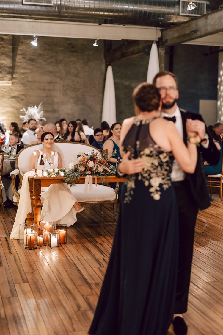 Bride watching son/parent dance inside industrial-style venue.
