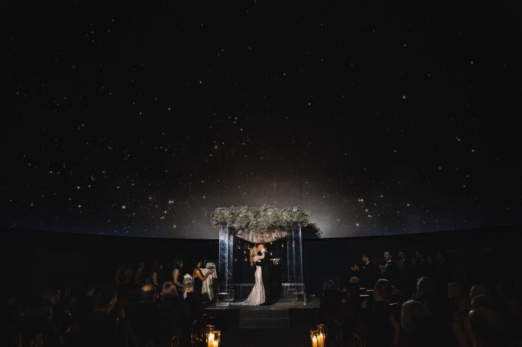 Bride and groom kissing under a floral arch during a nighttime ceremony.