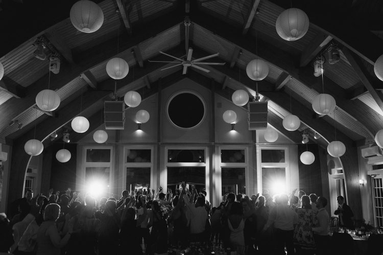 Crowded wedding dance floor inside vaulted hall with hanging lantern lights.