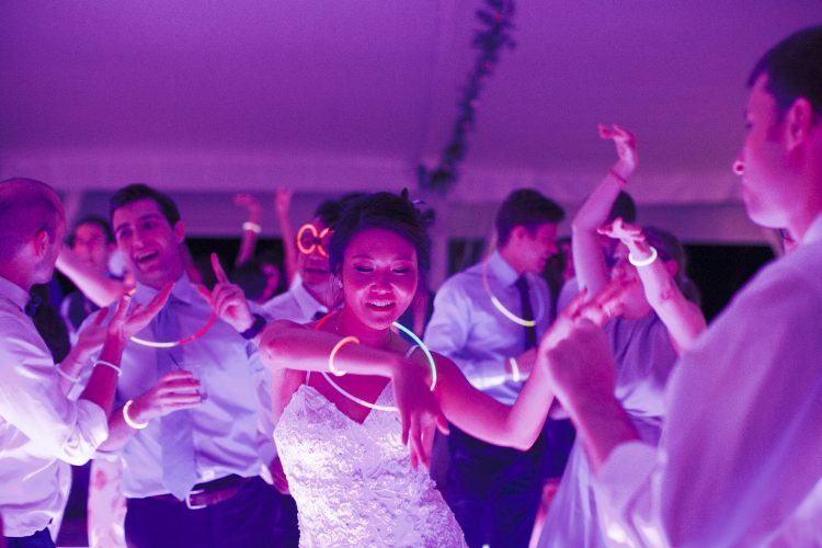 Bride dancing with guests wearing glow necklaces on colorful wedding dance floor.