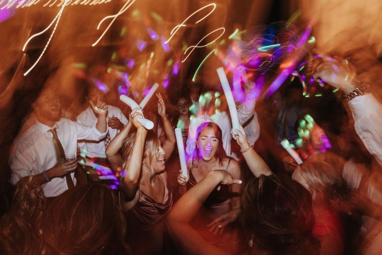 Wedding guests dancing with glow sticks on crowded reception dance floor.
