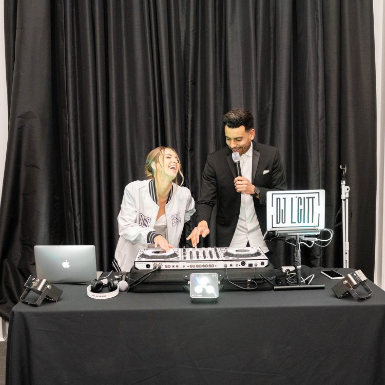 Bride DJing beside wedding DJ speaking into microphone at reception booth.