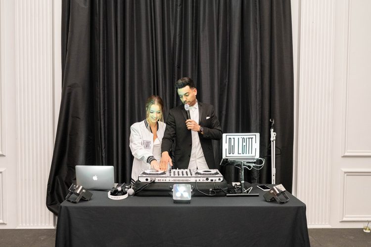 Bride DJing beside wedding DJ at reception booth.