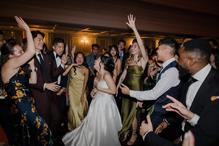 Bride dancing with cheering guests on crowded wedding reception dance floor.