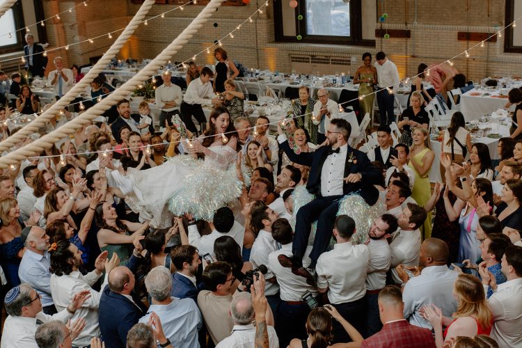 Bride and groom lifted in chairs during a lively wedding celebration.
