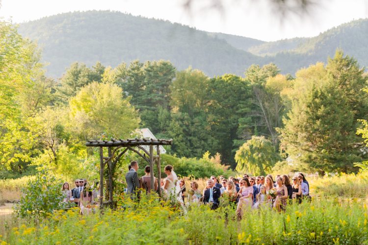 Outdoor wedding ceremony with guests seated in a scenic meadow.
