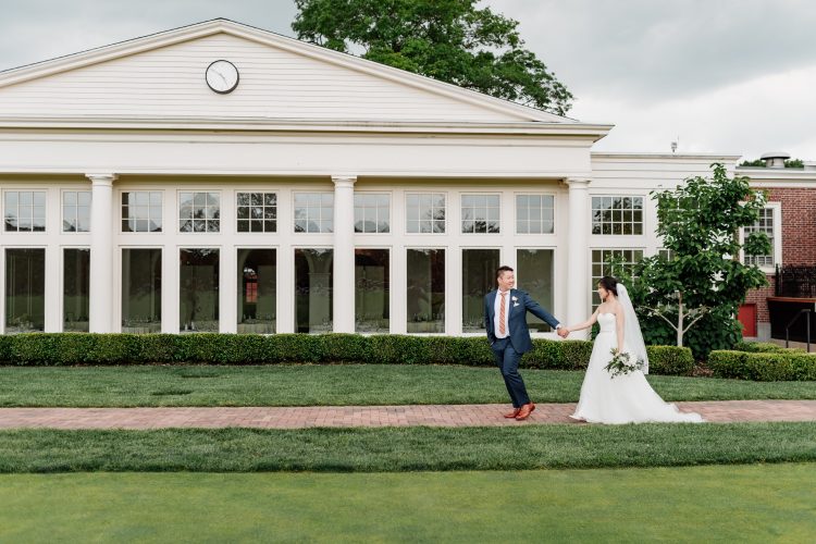 Bride and groom walking hand in hand in front of a white building at Philadelphia Cricket Club