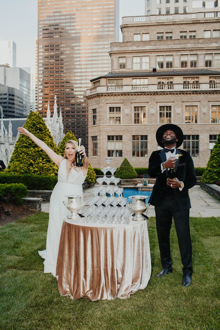 Bride and groom celebrating beside champagne tower outdoors with city skyline backdrop during wedding reception at 620 Loft & Garden