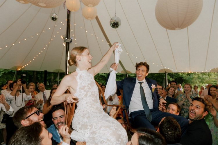 Bride and groom lifted by guests during energetic wedding reception under tented ceiling lights