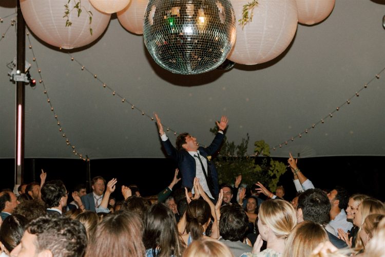 Groom lifted above wedding guests beneath disco ball during tented reception celebration