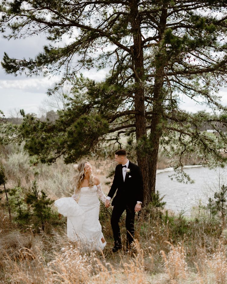 Bride and groom holding hands beneath pine trees beside a lake at Renault Winery
