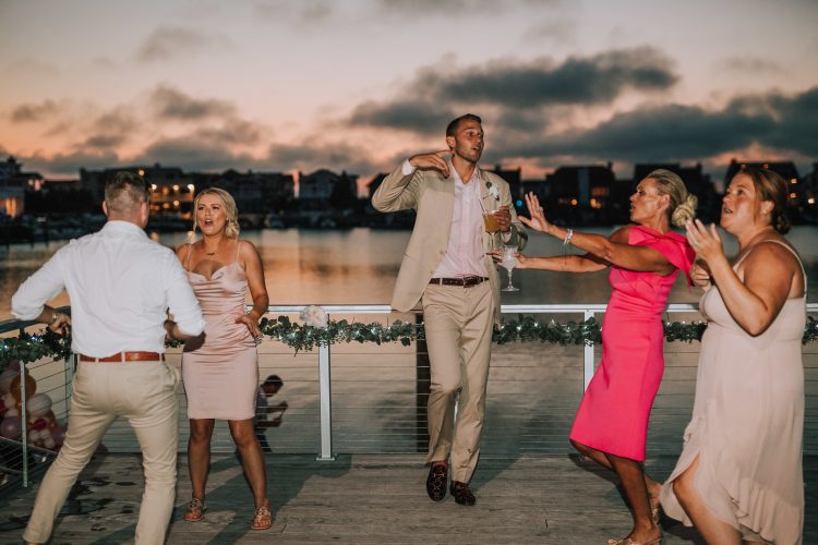 Wedding guests dancing on waterfront terrace during sunset reception.